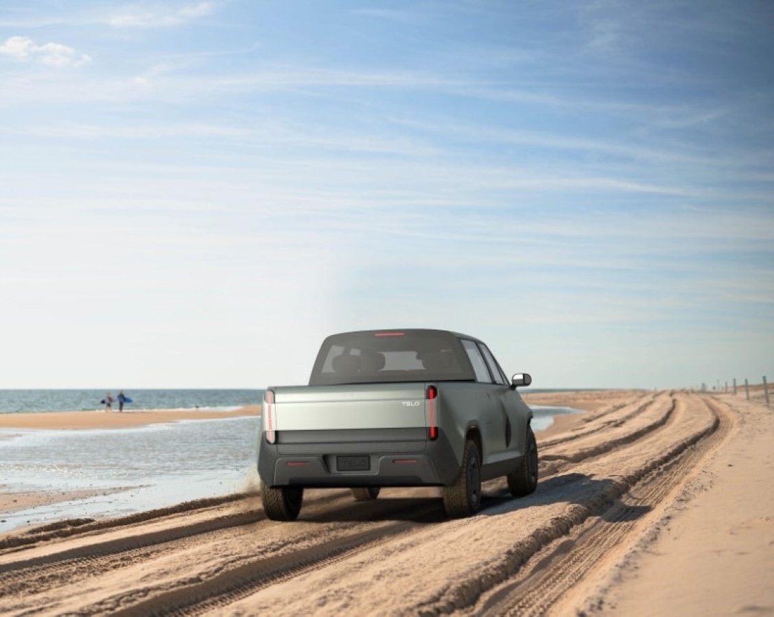 TELO truck from behind driving on the beach with surfers walking in the distnace.