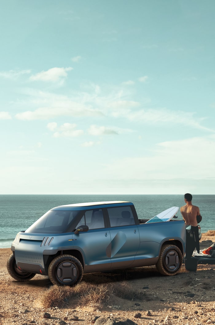 Blue TELO truck on a beach with a surfboard in the bed and a surfer looking at the ocean.